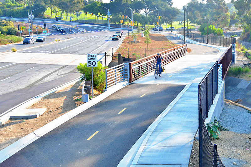 Southward view of Pershing Drive with a person riding a bike on a separated bikeway over a bridge. Balboa Park Golf Course is in the background showing the intersection of 26th Street and Florida Drive.