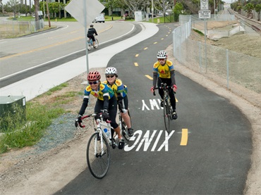 A roadway sign identifies the Bayshore Bikeway, carrying three cyclists alongside railroad tracks, separate from the sidewalk and the road. 