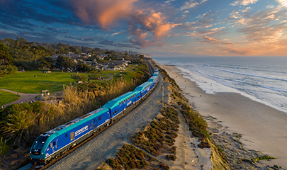 A Coaster train is traveling northbound near Seagrove Park. The train is traveling very close in-between a residential area and the beach cliffs. The beach and coastline is lit up from the blue and pink cloudy sky during dusk.