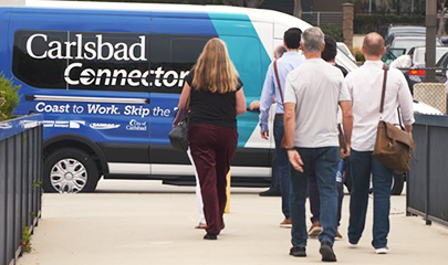 A group of people are walking on a sidewalk towards a blue and white small shuttle with the text "Carlsbad Connector" on the side of the vehicle.
