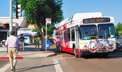 An MTS bus 30 is pulling away from a bus stop at an intersection with a market and Chinese restaurant in the background. People are walking near by. The bus has bikes stored on the front and electronic sign that says it is in route for UTC/VA Hospital via Pacific Beach.